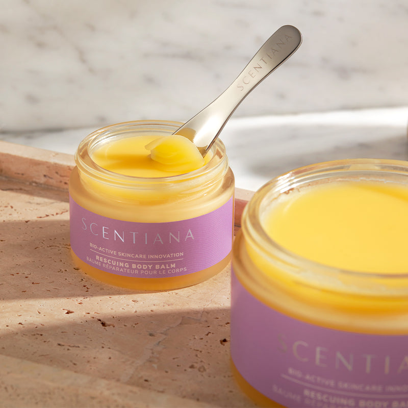 Two jars of Scentiana body balm on a concrete surface with a spatula inside one jar.